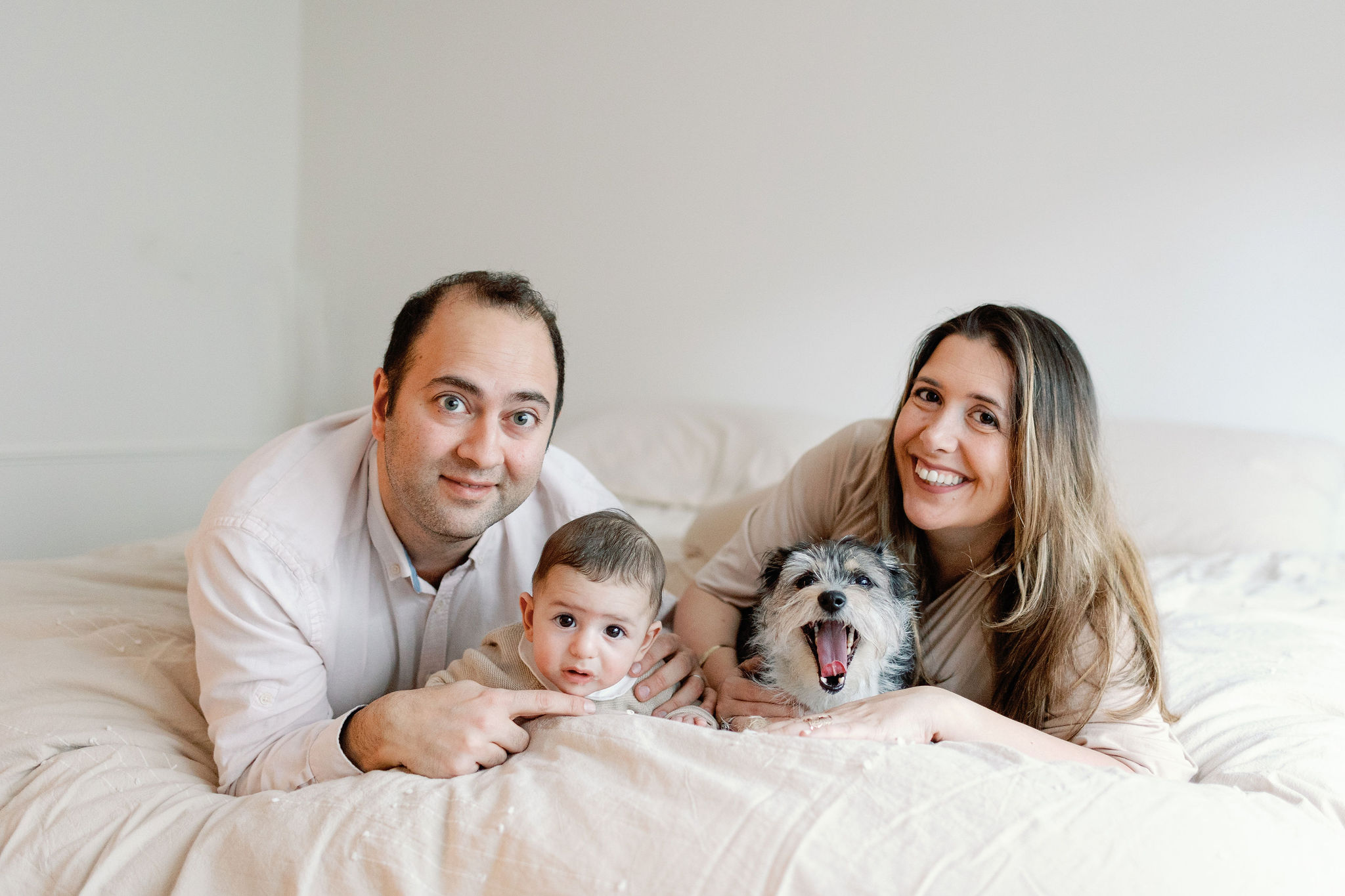 A couple, their newborn and their dog all stare forward and smile, while laying on a bed.