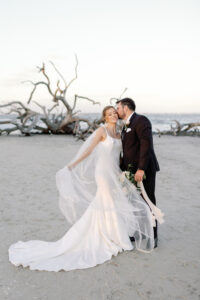The bride holds dress out on Driftwood beach while groom kisses her.